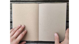 Zwei Hände halten ein leeres Notizbuch aus braunem Papier auf einer Holzoberfläche offen.