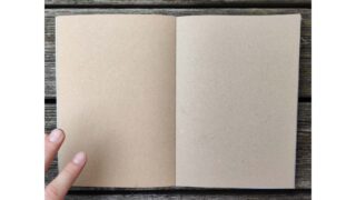 Ein offenes, leeres Notizbuch mit Seiten aus braunem Kraftpapier liegt auf einer Holzoberfläche; die linke Hand einer Person hält die Ecke der linken Seite.