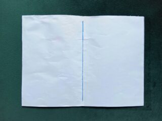 Ein offenes Heft aus weißem Papier mit einer sichtbaren blauen Fadenbindung in der Mitte, auf einer dunkelgrünen Oberfläche platziert.
