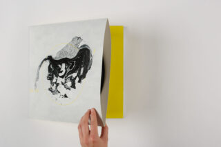 Eine Hand zieht eine Vinyl-Schallplatte teilweise aus einer quadratischen Hülle mit abstraktem schwarz-grauem Motiv auf weißem Hintergrund; die Innenseite der Hülle ist gelb.