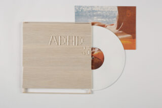 Eine weiße Vinyl-Schallplatte gleitet teilweise aus einer hellen Holzhülle mit der Prägung „ACHE 5“, im Hintergrund ist auf dem Albumcover eine entspannte, sonnenbeschienene Szene zu sehen.