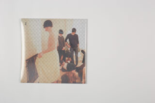 Ein quadratisches Blatt Papier mit dem Bild mehrerer Personen in einem Innenbereich, einige in Schwarz und eine in Weiß gekleidet, ist etwas links von der Mitte auf einem schlichten weißen Hintergrund platziert.