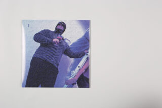 Ein aus niedriger Perspektive aufgenommenes Foto einer Person in einem gestreiften Pullover und einer Mütze, die neben einer anderen Person steht, die eine Gitarre hält, gedruckt auf einem weißen Hintergrund.