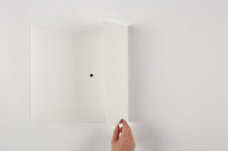 Eine Hand hebt die Kante eines weißen Ordners oder Umschlags an, in dessen Mitte sich auf einer schlichten weißen Oberfläche ein kleiner schwarzer Punkt befindet.