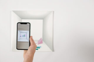 Eine Hand hält ein Smartphone, das eine Augmented-Reality-Ansicht bunter Formen über einem weißen quadratischen Kunstwerk an einer Wand zeigt.