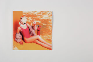 Eine Person in einem roten Badeanzug und einer roten Jacke sitzt vor einem orange marmorierten Hintergrund, trägt eine Pferdemaske und hält ein Getränk neben einem roten Feuerlöscher.