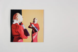 Ein Foto zeigt eine Person in einem roten Kleid und einer Fischmaske, die eine große Flasche hält und vor einem schlichten Hintergrund ihr Spiegelbild betrachtet.