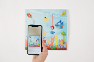 Eine Hand hält ein Smartphone und macht ein Foto von einem farbenfrohen Gemälde mit Unterwassermotiven, das Fische und Pflanzen auf einer weißen Wand zeigt.