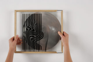 Eine Person hält einen quadratischen Holzrahmen mit einer Glasplatte und einer transparenten Vinyl-Schallplatte mit schwarzen Wellen- und vertikalen Linienmustern.