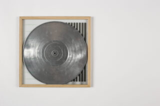 Eine Metallscheibe, die einer Vinyl-Schallplatte ähnelt, wird in einem Holzrahmen vor einem weißen Hintergrund präsentiert.