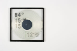 Auf einer transparenten Scheibe mit Rahmen sind die numerischen Koordinaten in Schwarz und Grün zu sehen: 64° 15'N und 12°W auf einem schlichten weißen Hintergrund.