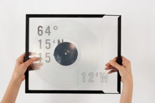 Zwei Hände halten eine gerahmte transparente Folie mit konzentrischen Kreisen und den darauf aufgedruckten Koordinaten „64° 15'N 15 12'W“.