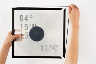 Zwei Hände halten einen transparenten quadratischen Rahmen mit einer Schallplatte und fettgedruckten Koordinaten: 64°15'N 25°12'W vor einem weißen Hintergrund.