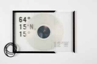 Ein transparentes rechteckiges Objekt mit schwarzem Rahmen, konzentrischen Kreisen in der Mitte, schwarzer Kordel auf der linken Seite und den auf der Oberfläche aufgedruckten Koordinaten „64°15’N, 15°12’W“.