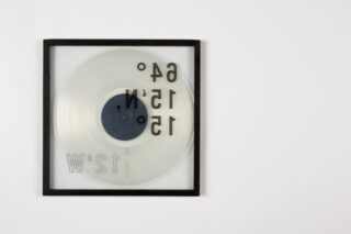 Ein quadratischer Rahmen, der eine transparente runde Scheibe mit umgekehrt angeordneten schwarzen und weißen Zahlen und Buchstaben auf weißem Hintergrund enthält.