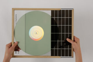 Zwei Hände halten ein gerahmtes Kunstwerk mit einer großen, durchscheinenden runden Scheibe mit einem holografischen Zentrum, über dem sich ein schwarz-weißes Gittermuster und teilweise sichtbarer Text befinden.