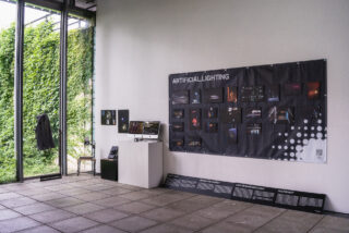 Ausstellungsraum mit einer Präsentation über künstliche Beleuchtung, mit Fotos, Informationstafeln, einem Computer und einem Mantel, der an Fenstern mit Blick auf eine grüne Efeuwand hängt.