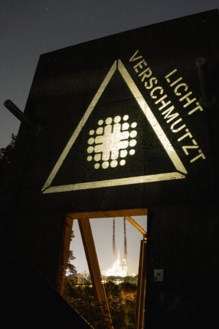 Ein dreieckiges Schild mit der Aufschrift „LICHT VERSCHMUTZT“ ist nachts beleuchtet; durch eine rechteckige Öffnung unter dem Schild ist ein weit entferntes beleuchtetes Gebäude sichtbar.