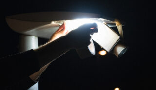 Eine Person justiert oder repariert nachts mit der Hand eine Überwachungskamera und eine Leuchte, wobei ein helles Licht die Szene beleuchtet.