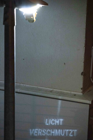 Eine beleuchtete Straßenlaterne projiziert die deutschen Worte „LICHT VERSCHMUTZT“ auf eine Wand und macht so auf das Problem der Lichtverschmutzung aufmerksam.