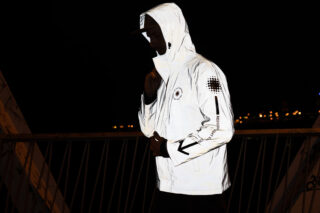 Eine Person, die eine reflektierende weiße Jacke mit Kapuze trägt, steht nachts im Freien, im Hintergrund sind die Lichter der Stadt verschwommen.