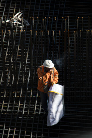 Ein Bauarbeiter in einer orangefarbenen Jacke und einem weißen Helm steht zwischen Stahlbewehrungsstäben, hält ein großes Laken oder einen Sack und bedeckt seinen Mund.