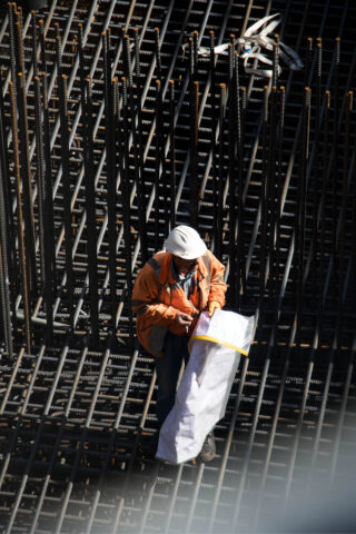 Ein Bauarbeiter mit Schutzhelm und orangefarbener Jacke untersucht einen großen Bauplan, während er auf einer Baustelle zwischen vertikalen und horizontalen Stahlbewehrungen steht.