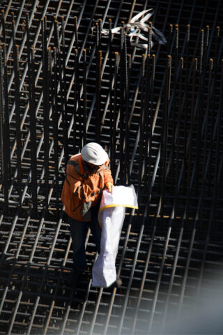 Ein Bauarbeiter mit Schutzhelm und orangefarbener Jacke untersucht auf einer Baustelle die Baupläne einer Bewehrungskonstruktion.