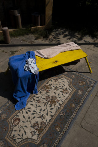 Eine gelbe Holzbank mit einem beige-blauen Handtuch darüber, neben einem gemusterten Teppich auf einer Steinterrasse. Die Szene spielt im Freien bei teilweisem Sonnenlicht.