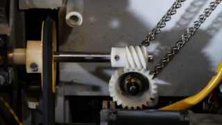 Nahaufnahme eines mechanischen Systems mit einer Metallwelle, einem Kunststoffschneckengetriebe, einem Zahnrad und einer Gliederkette im Inneren einer Maschine.