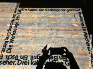 Die Hände einer Person, die ein Telefon hält, werfen einen Schatten auf einen Holzboden, an dessen Rändern deutscher Text und ein Bildnachweis projiziert werden.