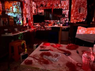 Ein schwach beleuchteter Raum mit roten Schnüren und Garn, die auf Tischen und Oberflächen verstreut sind und durch rote Lichtprojektionen beleuchtet werden, die die Wände und Möbel bedecken.