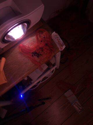 Ein Projektor wirft Licht auf einen Tisch mit verheddertem orangefarbenem Seil, einer kleinen Notiz mit der Aufschrift „BÜHNE“ und einigen verstreuten Gegenständen auf einem Holzboden.