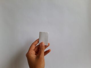 Eine Hand hält ein kleines, quadratisches Stück weißes Papier vor einem hellgrauen Hintergrund.