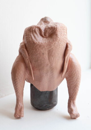 Ein rohes, gerupftes ganzes Huhn steht aufrecht auf einem dunklen zylindrischen Objekt vor einem hellen Hintergrund.