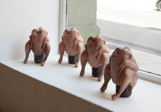 Vier rohe, gerupfte Hühner liegen kopfüber auf schwarzen zylindrischen Objekten auf einem Fensterbrett. Durch das Fenster ist eine Straßenszene zu sehen.