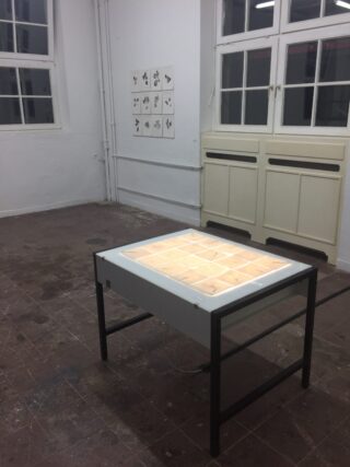 Ein beleuchteter Ausstellungstisch mit Objekten darin steht in der Mitte eines leeren Raums mit gefliestem Boden und mehreren Schwarzweißzeichnungen an der Wand.