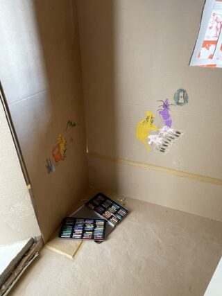 Eine Zimmerecke mit braunen Pappwänden, Kinderzeichnungen an der Wand und offenen Pastellkästen auf dem Boden.