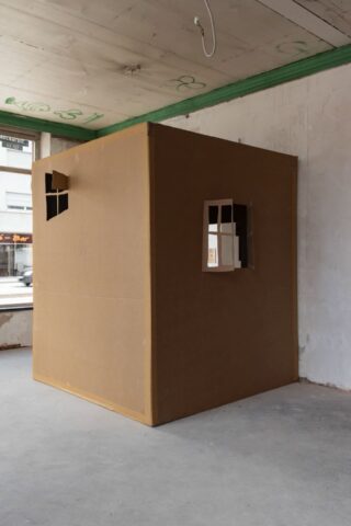 Eine große, braune, kastenförmige Struktur mit zwei kleinen Fenstern steht in einem leeren, unfertigen Raum mit Betonböden und freiliegenden Deckenkabeln.