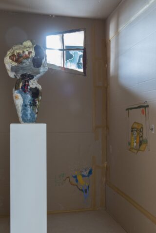 Eine bemalte Skulptur steht auf einem weißen Sockel in einem Raum mit Pappwänden, kindlichen Zeichnungen und Licht, das durch ein kleines Fenster strömt.