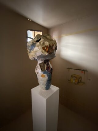 Eine geformte Figur mit offenem Mund und gezackten Zähnen steht auf einem weißen Sockel in einem schwach beleuchteten Raum mit einem kleinen Fenster und einem Kunstwerk an der Wand.