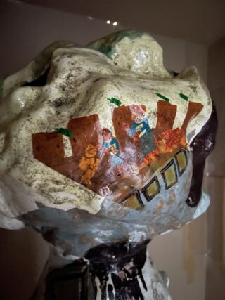 Eine Nahaufnahme einer bemalten Keramikskulptur, die eine teilweise verblasste Szene von Menschen vor braunen Gebäuden auf einer rauen, strukturierten Oberfläche zeigt.