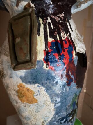 Nahaufnahme einer bemalten Keramikskulptur mit abstrakten Mustern, einem rechteckigen hervorstehenden Element und verschiedenen Farben, darunter Blau, Rot, Braun und Gelb.