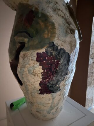 Eine strukturierte Keramikvase mit abstrakten gemalten Mustern in Grün, Rot und Schwarz, platziert auf einer weißen Oberfläche neben einem grünen Universalmesser.