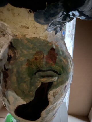 Eine bemalte Keramikoberfläche mit zwei kleinen menschlichen Figuren in Grün- und Brauntönen, darunter eine große dunkle Form und ungleichmäßigen Farbmustern.