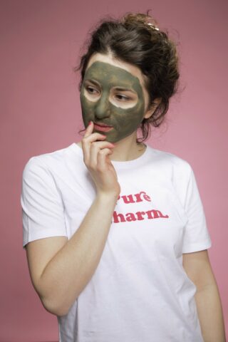 Eine Frau in einem weißen T-Shirt mit rotem Text steht vor einem rosa Hintergrund, hat eine grüne Gesichtsmaske auf dem Gesicht und berührt mit der Hand ihr Kinn.