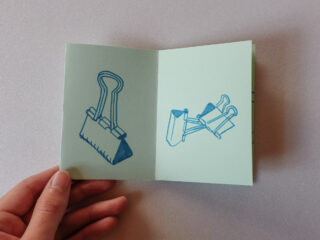 Eine Hand hält ein offenes Heft mit zwei blauen Strichzeichnungen von Heftklammern auf hellgrünem Papier.