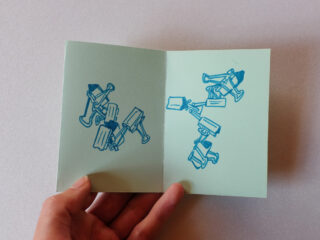 Eine Hand hält eine offene Karte mit blauen Abbildungen von Heftklammern, die auf beiden Seiten vor einem schlichten Hintergrund verstreut sind.