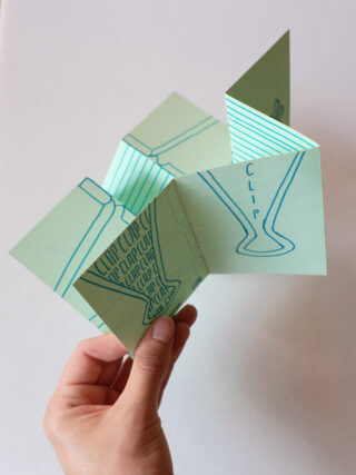 Eine Hand hält ein kunstvoll gefaltetes Papierkunstwerk mit geometrischen Formen und blauen Strichzeichnungen, darunter das Wort „CLIP“ und stilisierte Cliparts.