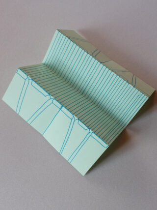 Ein gefaltetes Stück grünes Papier mit blauen Linien, die geometrische Muster bilden, auf einer flachen, hellen Oberfläche platziert.